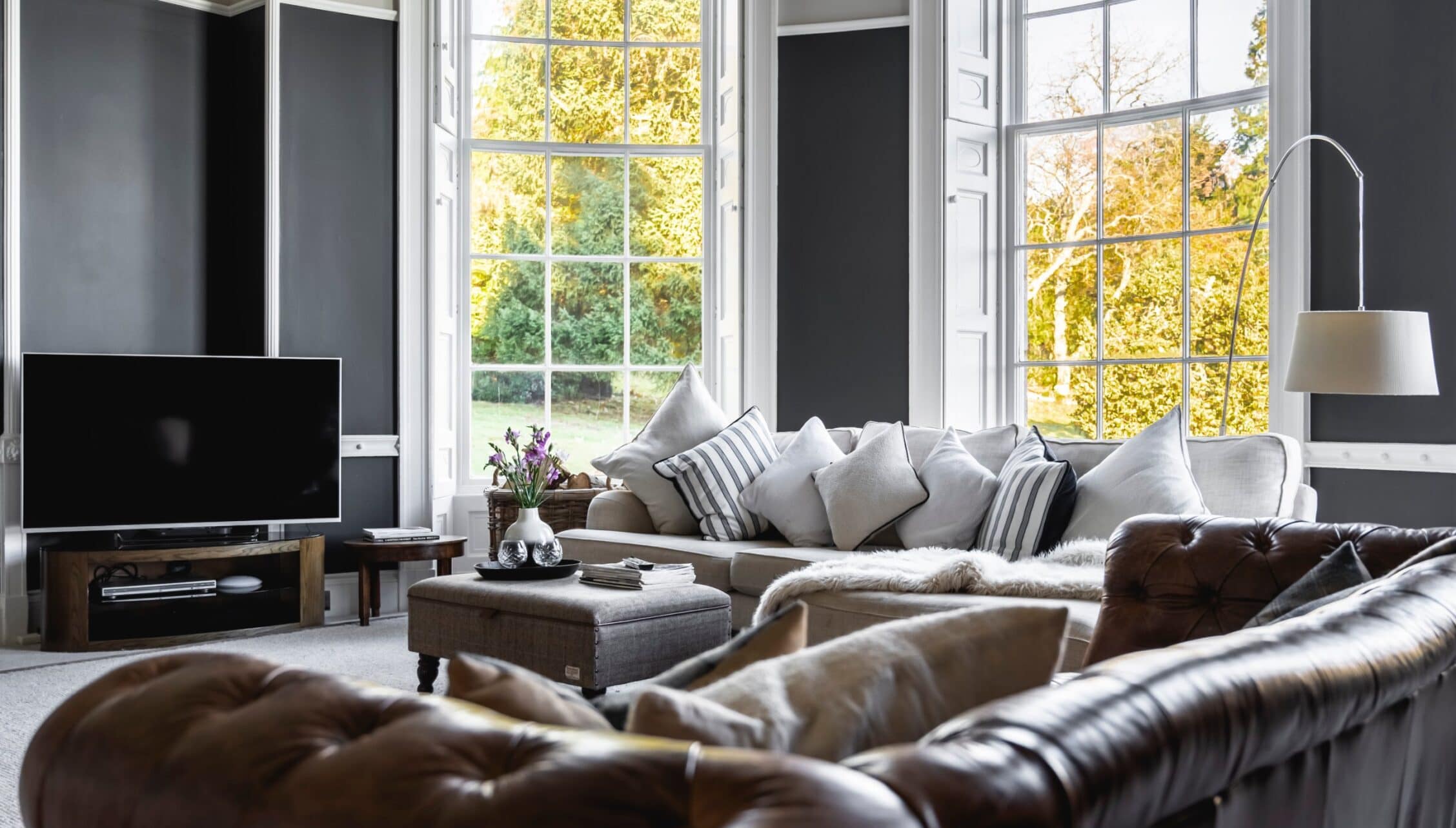 Manar House, Aberdeenshire – Spacious sitting room with grey upholstered sofa, leather armchair, and sash windows overlooking autumn trees.