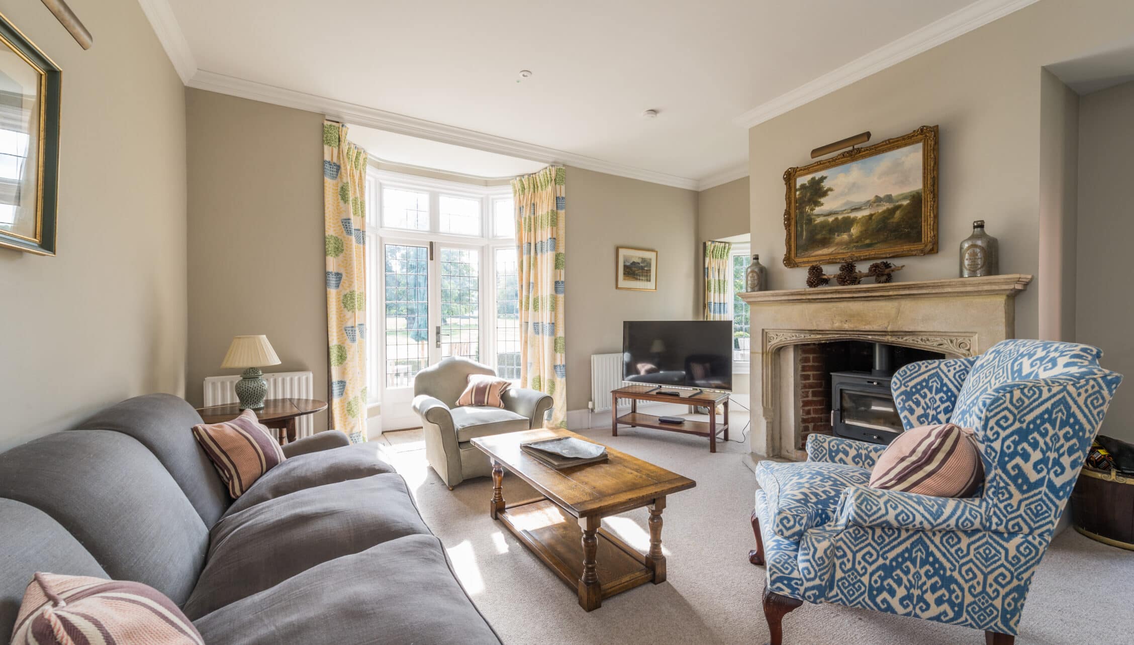 Nedging Hall, Suffolk – Spacious sitting room with bay windows, fireplace and patterned upholstered armchairs.