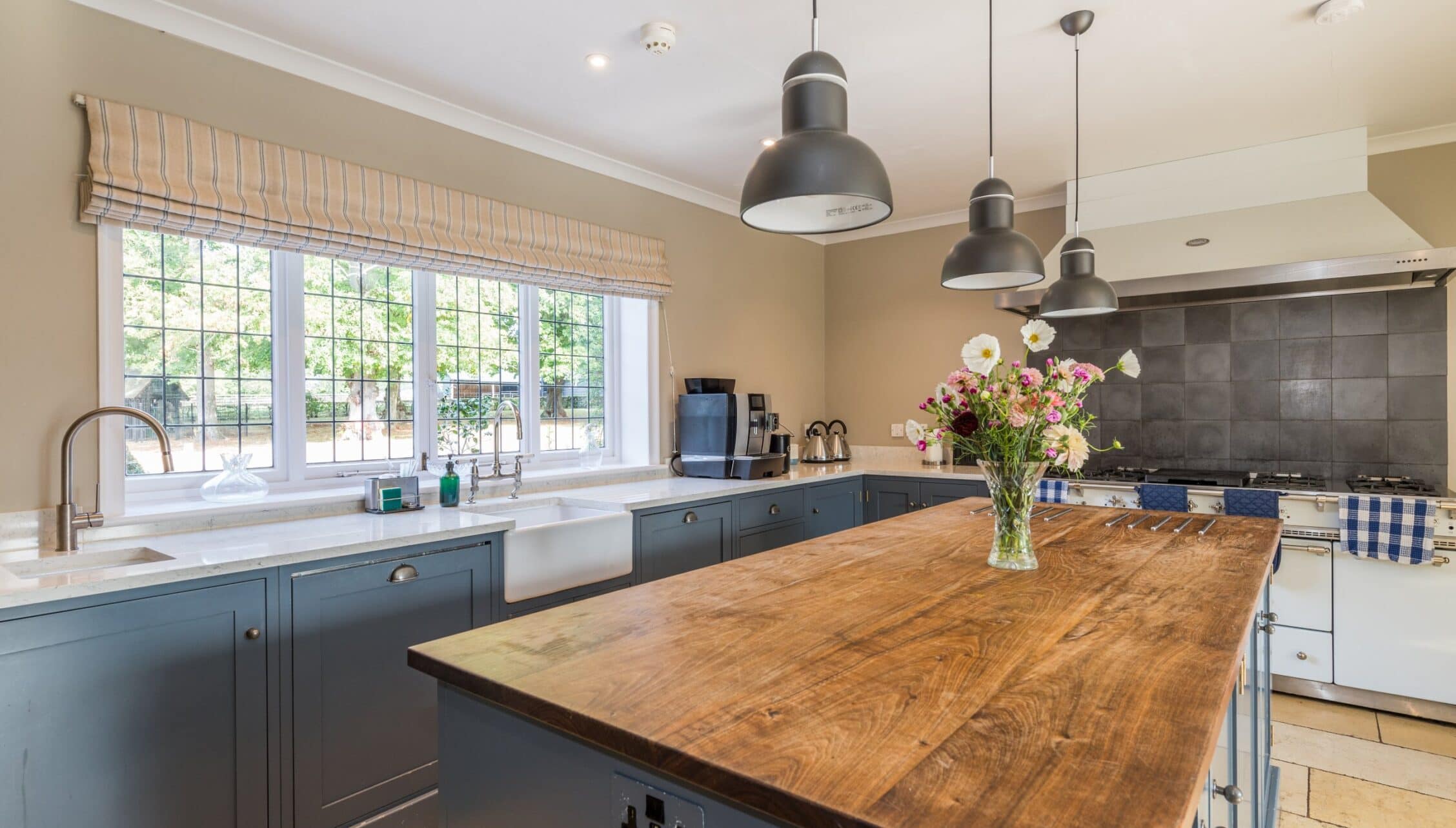 Nedging Hall, Suffolk – Kitchen with blue cabinetry, wooden island, and pendant lighting overlooking garden.