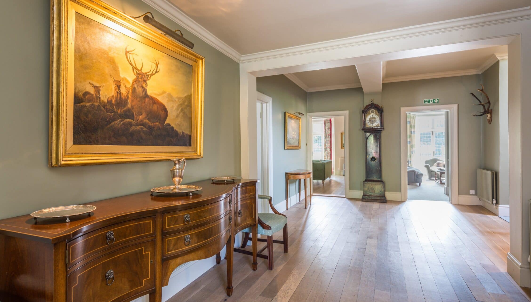 Nedging Hall, Suffolk – Hallway with inlaid wood dresser, gilt-framed stag painting and grandfather clock.