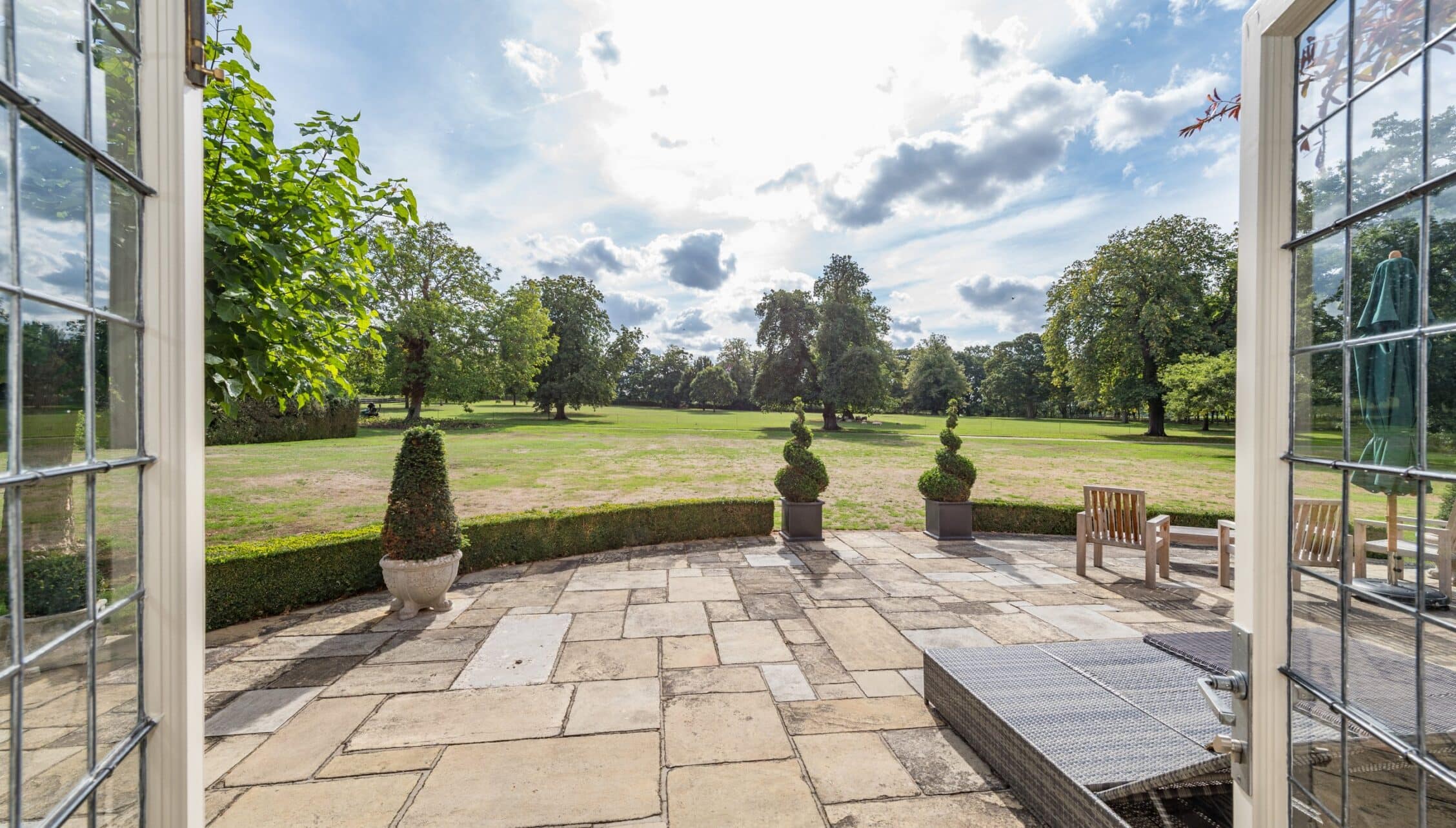 Nedging Hall, Suffolk – Stone patio with topiary planters overlooking parkland and mature trees.
