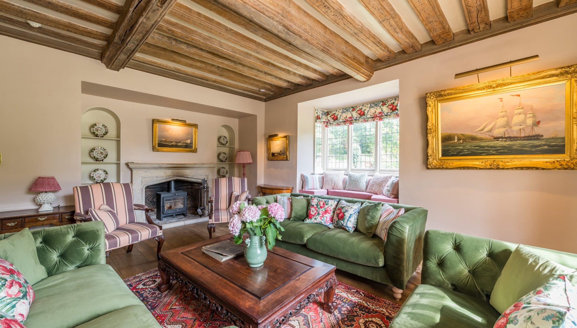 Nedging Hall, Suffolk – Beamed drawing room with green velvet sofas, wood stove and gilt-framed maritime paintings.