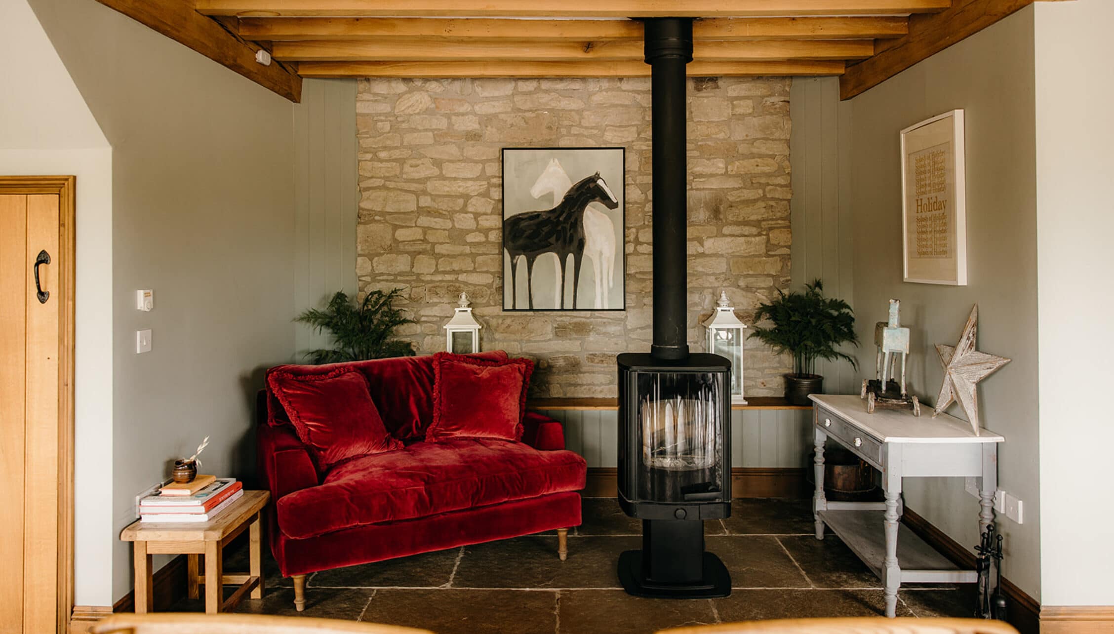 The Barns – Exclusive Hire, Herefordshire – Living room with red velvet sofa, wood-burning stove and exposed stone fireplace.