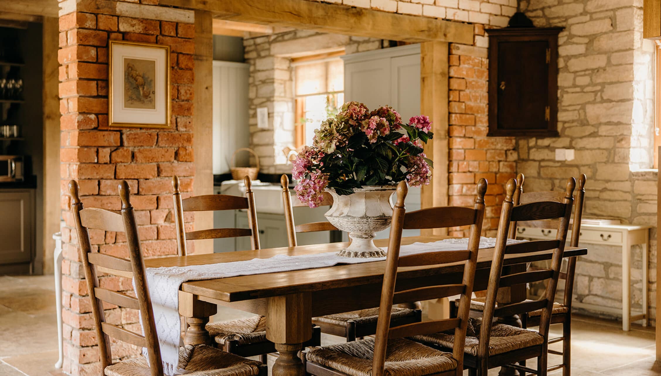 The Barns – Exclusive Hire, Herefordshire – Dining room with exposed brick walls, wooden ladder-back chairs, and pink flowering arrangement in urn.