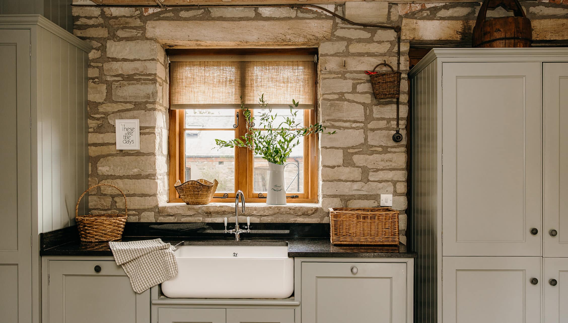 The Barns – Exclusive Hire, Herefordshire – Stone country kitchen with cream cabinetry, black worktops and ceramic farmhouse sink beneath wooden-framed.