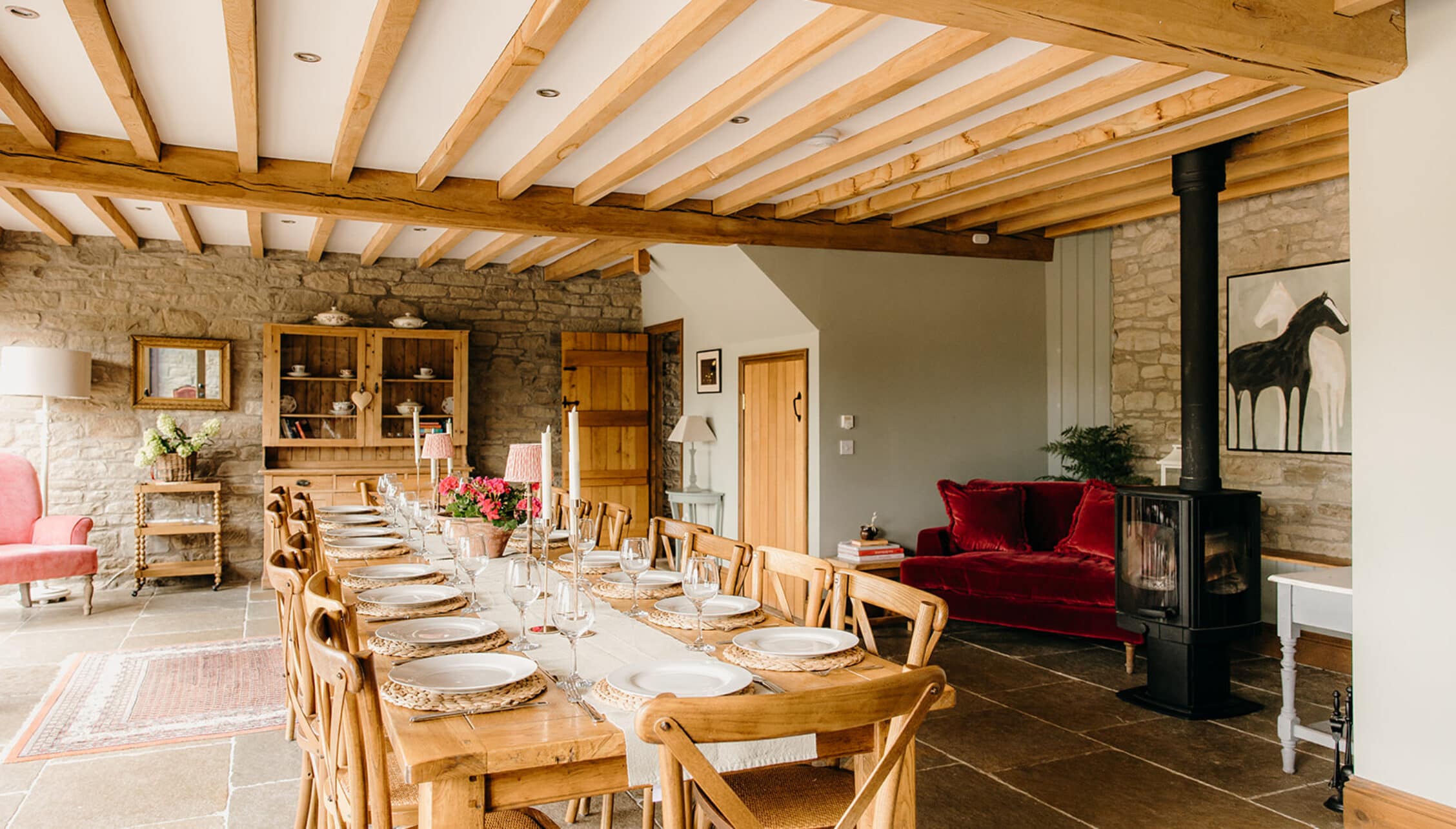 The Barns – Exclusive Hire, Herefordshire – Open-plan dining and sitting room with exposed wooden beams, stone walls, and wood-burning stove.