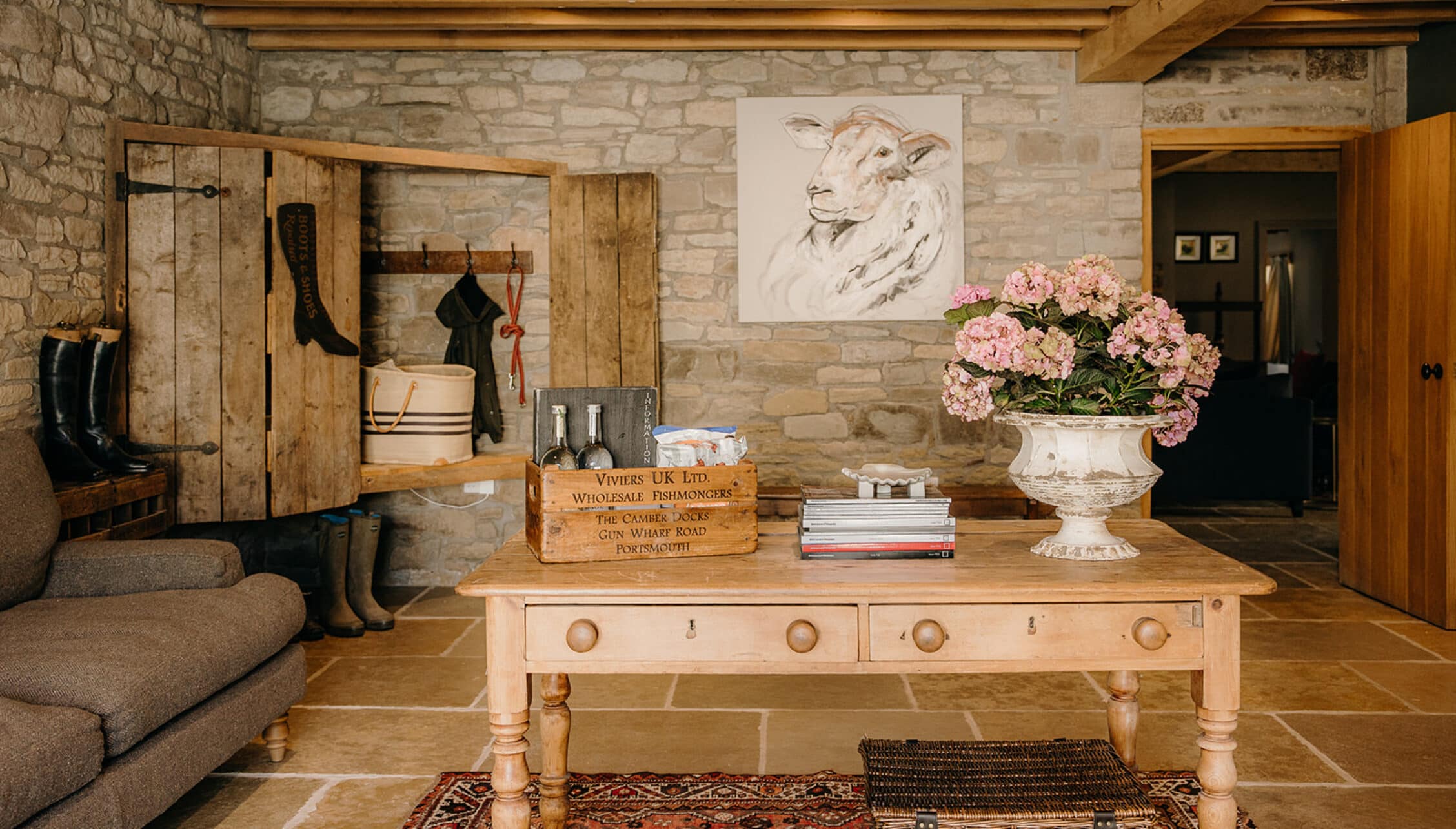 The Barns – Exclusive Hire, Herefordshire – Stone barn interior with wooden storage shelving, pink hydrangeas in urn, and wooden console table.