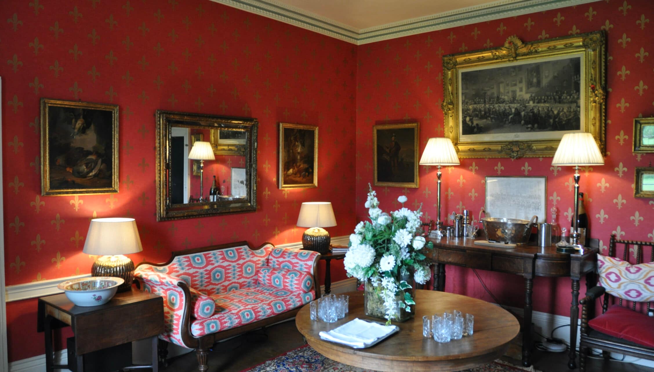 Wardhill Castle, Aberdeenshire – Red wallpapered drawing room with gilt-framed paintings, ornate mirror and round wooden table.