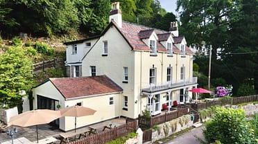 Holly Tree House – Cream-rendered country house with terracotta roof, patio seating area and wooded hillside setting.