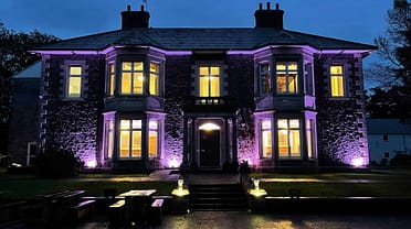 Plas Y Bryn – Stone country house exterior at dusk, illuminated by purple uplighting, with ivy-covered walls and sash windows.