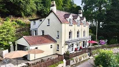 Holly Tree House – Cream-rendered country house with terracotta roof, patio seating area and wooded hillside setting.
