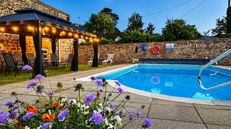 Brookside, Herefordshire – Swimming pool with stone boundary wall, lit pergola, and purple flowers.